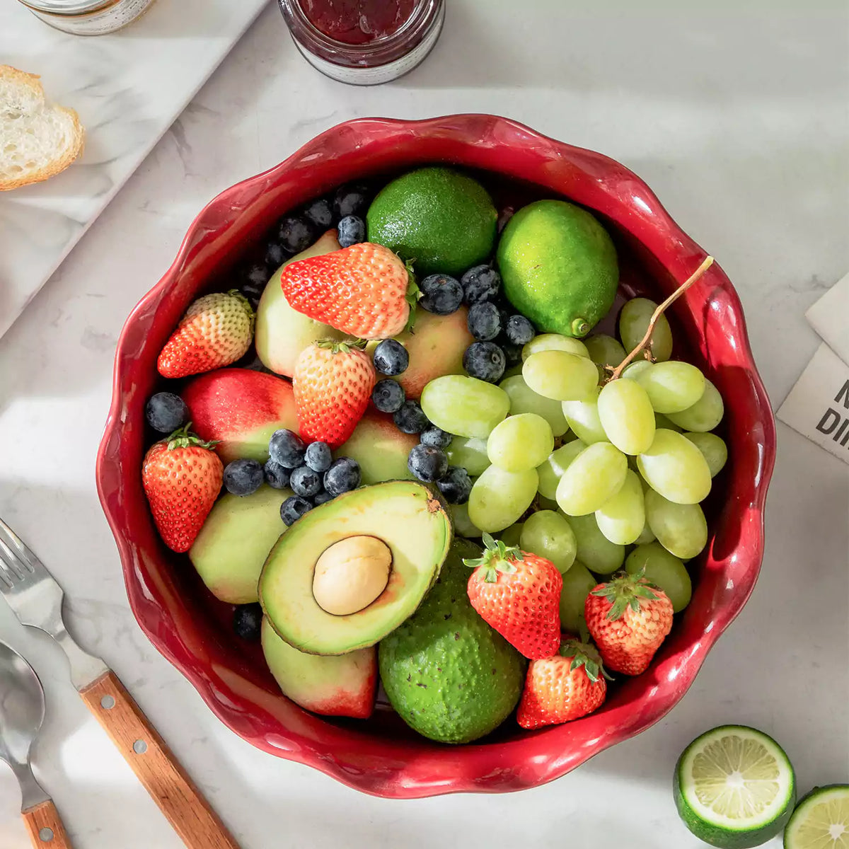 Cherry red fluted pie pan with mixed berries, avocado, grapes, jam, and lime on marble table.