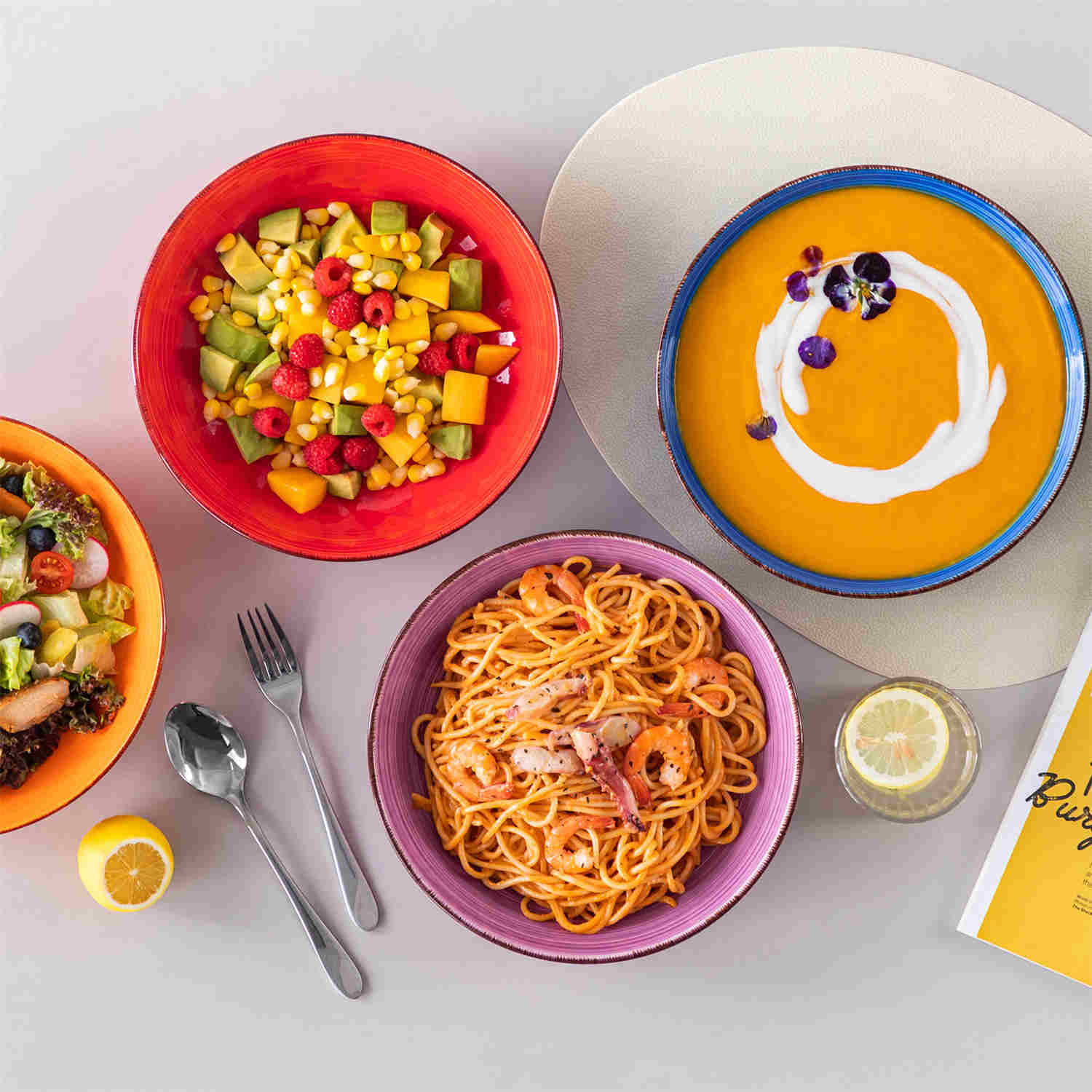 Multicolor Bonita large stoneware bowls set of 6 on a white table - each filled with pasta - pumpkin soup - and fruit salad - creating a bright and inviting table setting