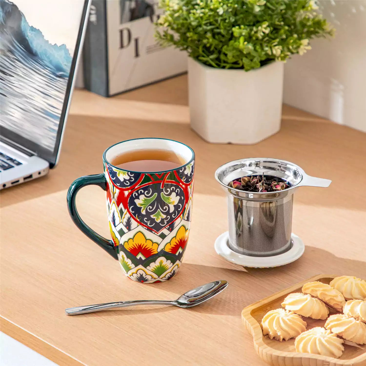 Lumi forest green porcelain tea mug with infuser and lid featuring vibrant Bohemian pattern on a wooden desk with laptop and plant