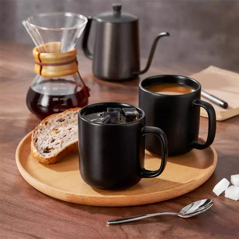Playa 16oz stoneware mugs in black reactive glaze on a rustic wooden café table with coffee maker and pastries