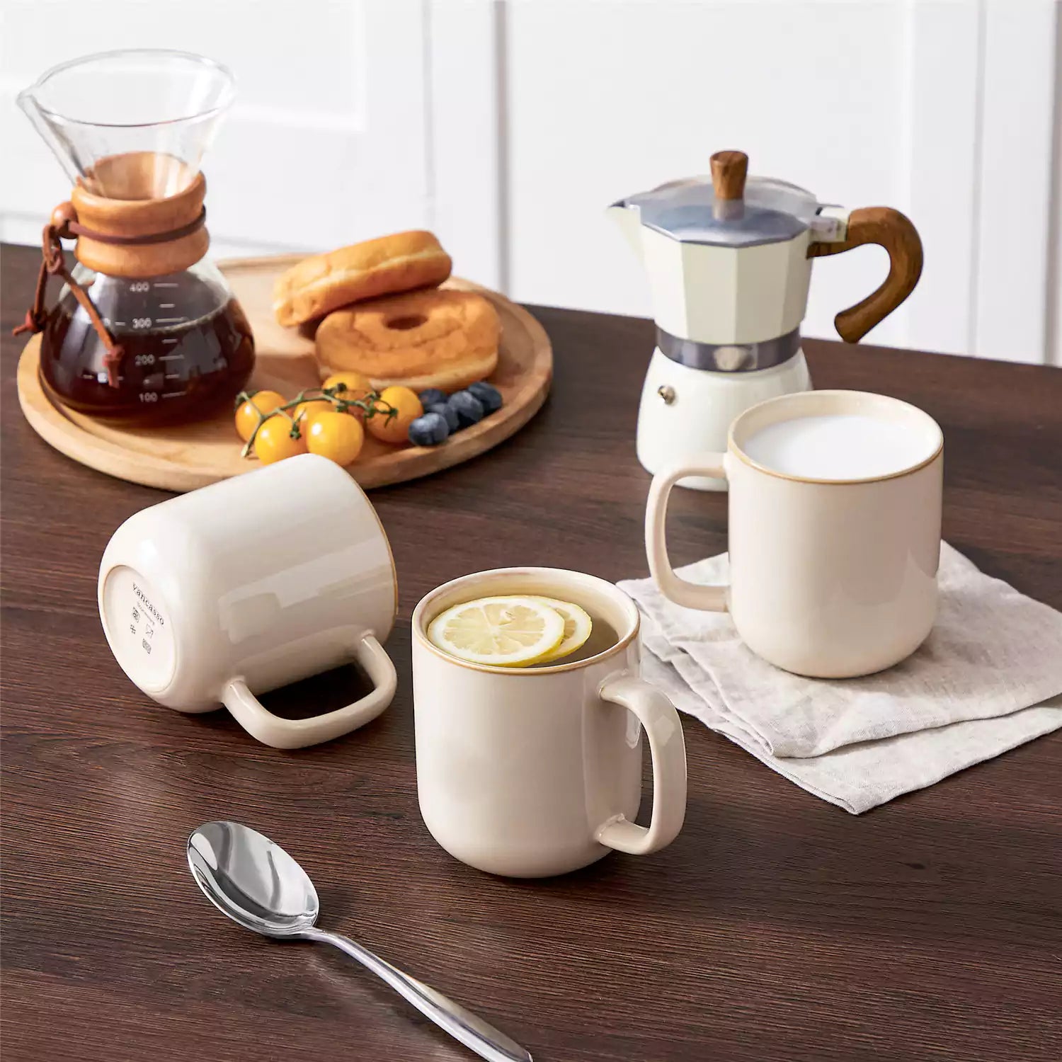 Playa 16oz stoneware mugs in beige reactive glaze on a rustic wooden café table with coffee maker and pastries