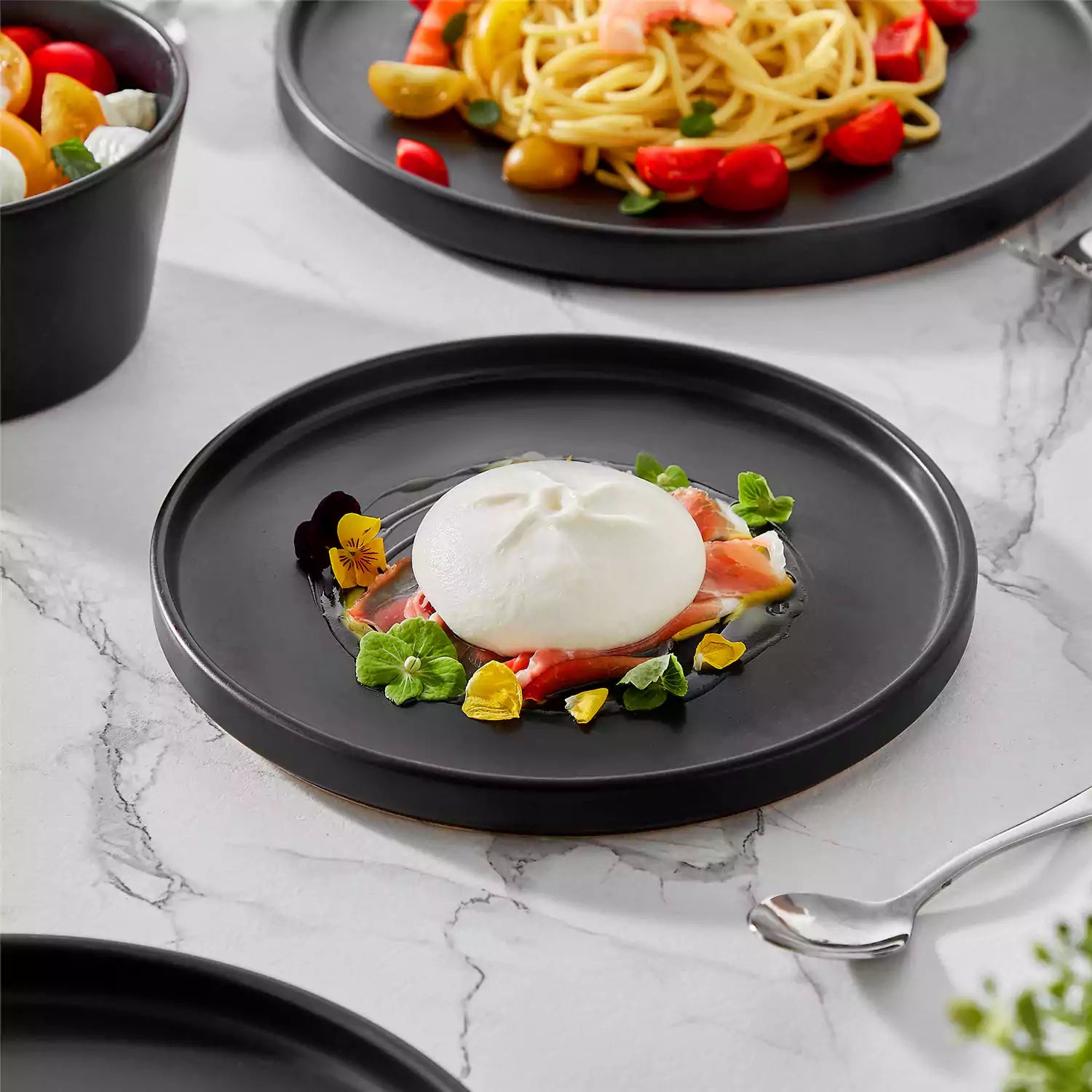 Modern black stoneware bowl and plate set displayed on a marble countertop - with a fresh salad blurred in the background - vancasso