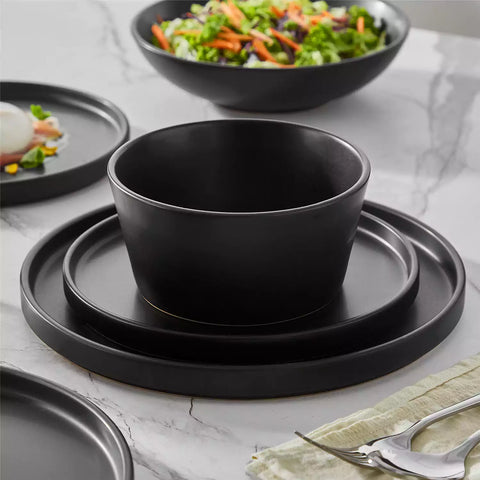 Modern black stoneware bowl and plate set displayed on a marble countertop - with a fresh salad blurred in the background - vancasso