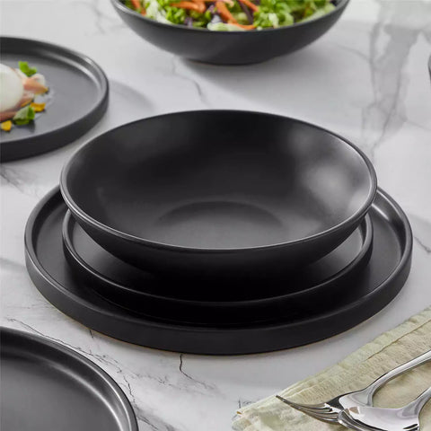 Modern black stoneware bowl and plate set displayed on a marble countertop - with a fresh salad blurred in the background - vancasso