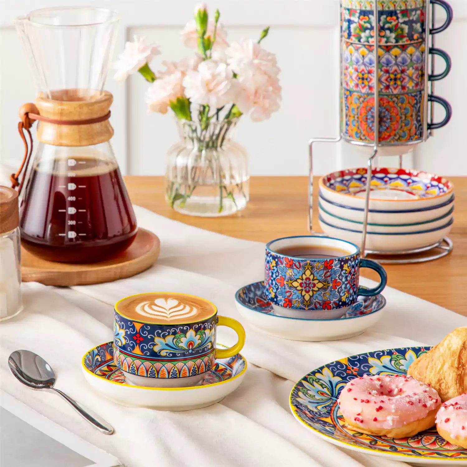 Simi 12 piece porcelain cafe set with 5oz flat white cups and saucers in vibrant boho chic floral patterns on a black metal storage rack with a magazine and napkin on a wooden table