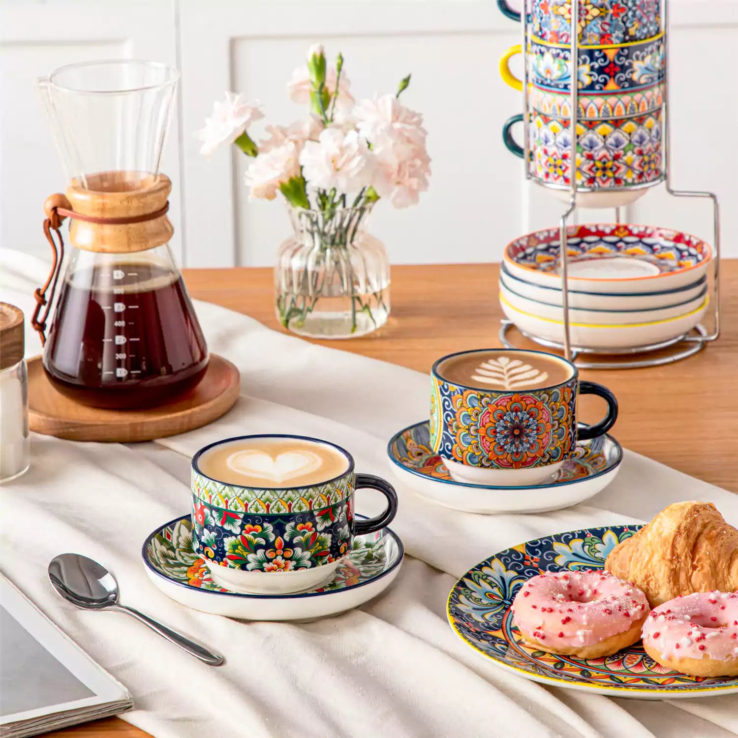 Simi 12 piece porcelain cafe set with 9oz flat white cups and saucers in vibrant boho chic floral patterns on a black metal storage rack with a magazine and napkin on a wooden table