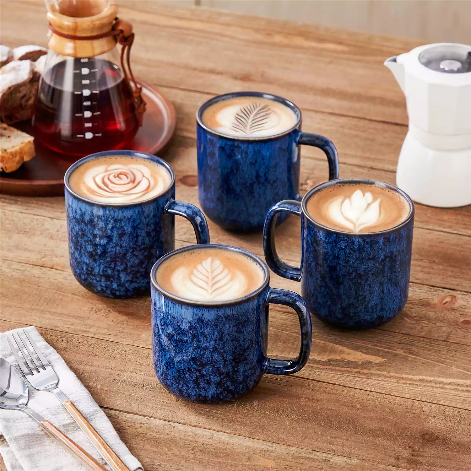 Four starry blue 16oz stoneware mugs with latte art on wooden table beside coffee brewing equipment
